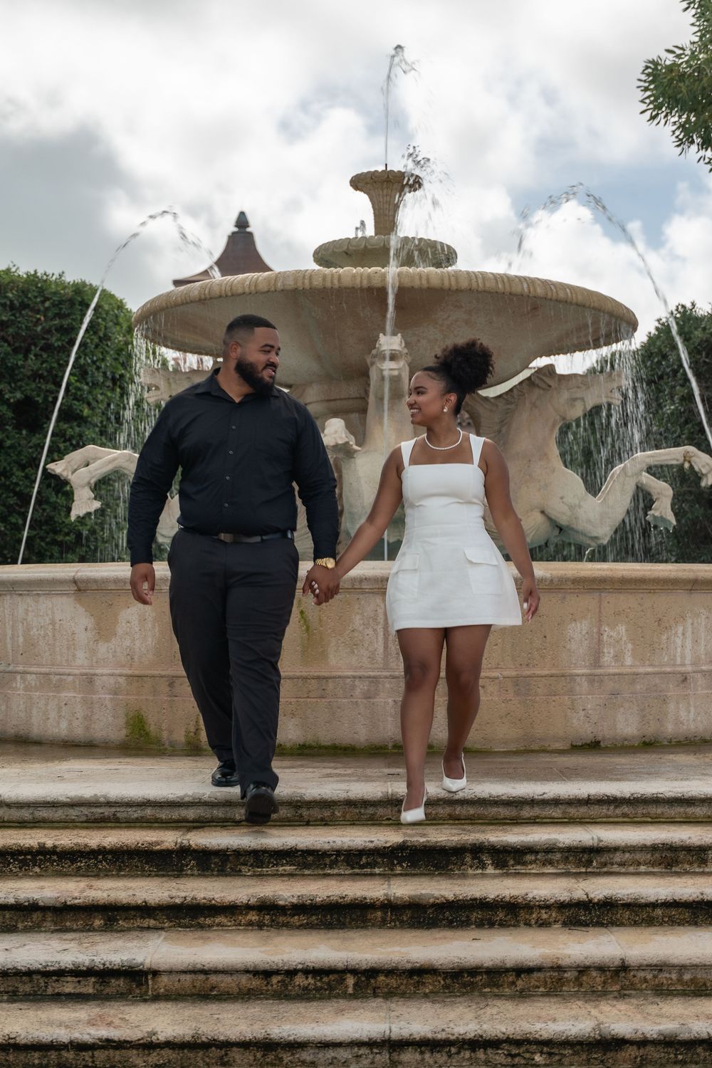 Couple holding hands walking down steps in front of an ornate stone fountain with horse sculptures outdoors.