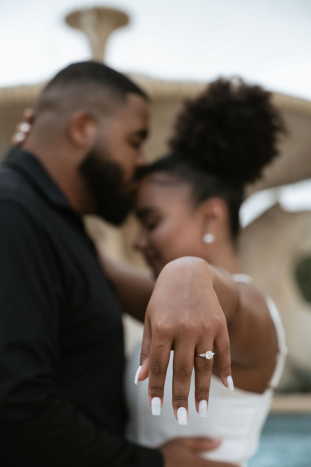 Couple sharing a romantic moment while woman displays her diamond engagement ring with white manicured nails.