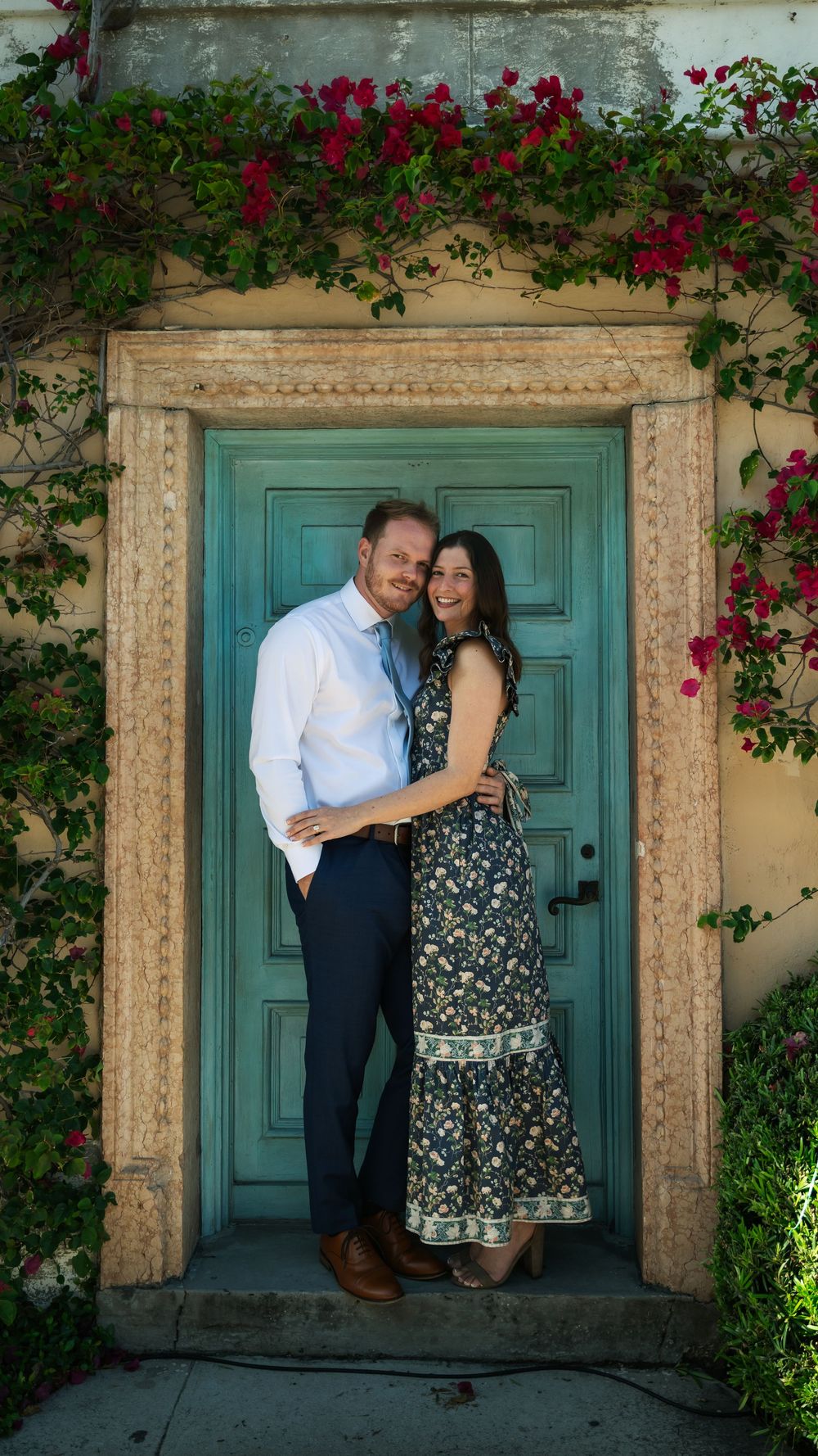 Couple smiling and embracing in a teal doorway framed by bougainvillea flowers on a stone wall.