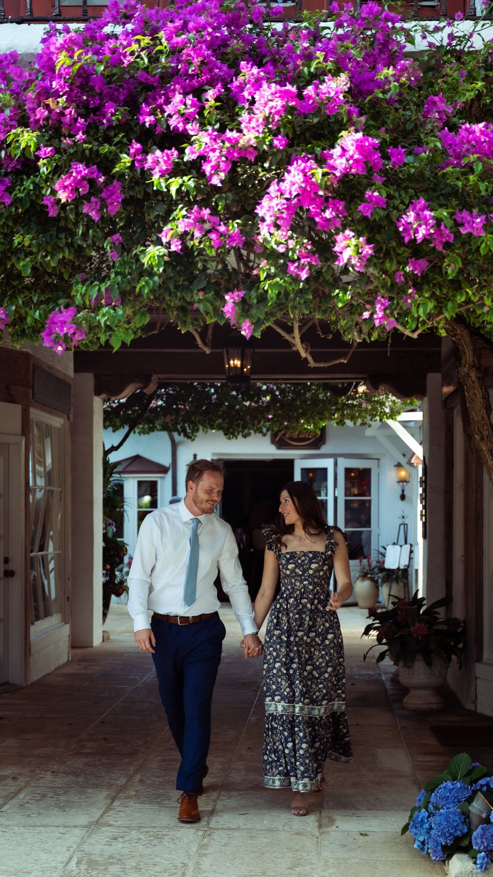 Couple walks hand-in-hand under vibrant purple bougainvillea archway at elegant venue.