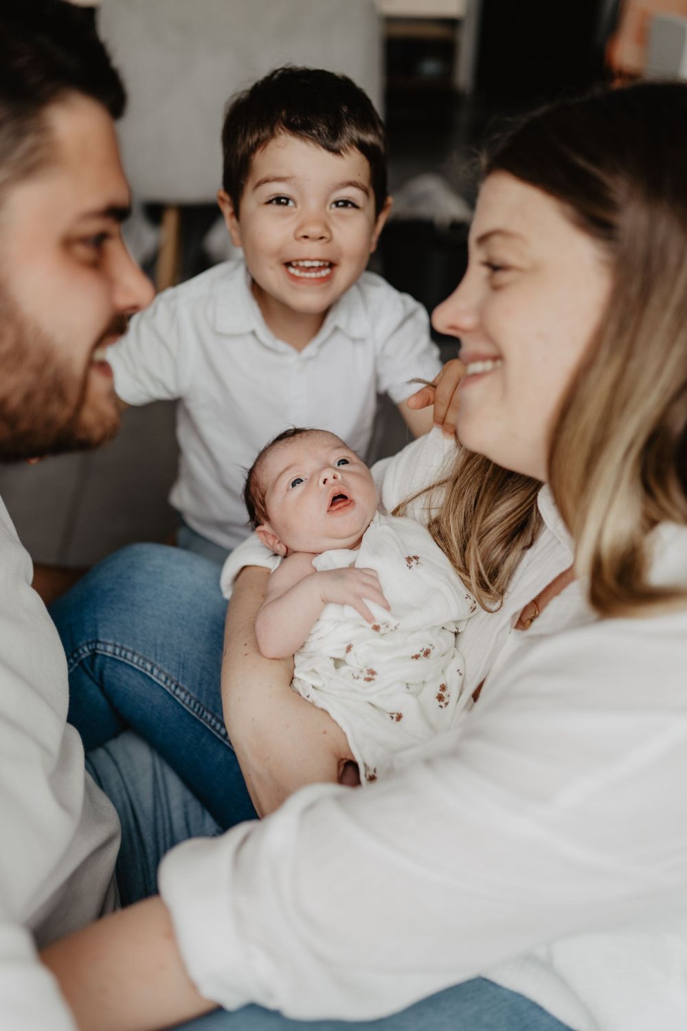 Un nouveau-né vêtu de blanc est tenu doucement dans les bras de ses parents avec son petit frère en arrière plan