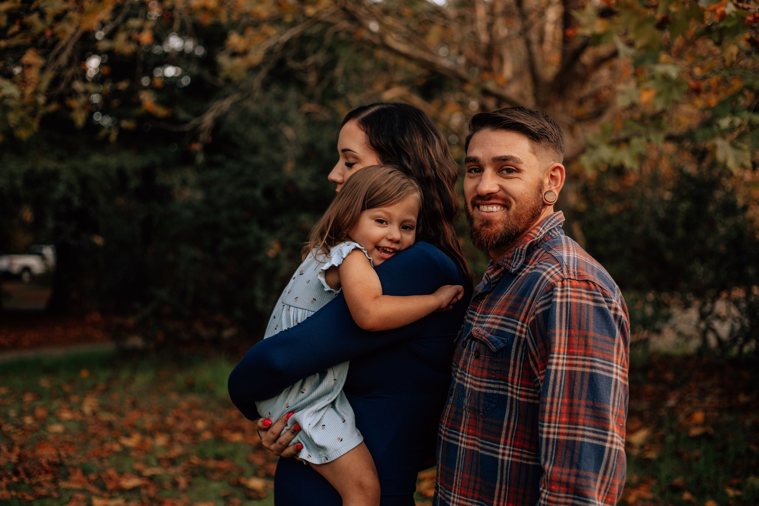A family shares a warm embrace while standing in a park during autumn with fallen leaves in the background.