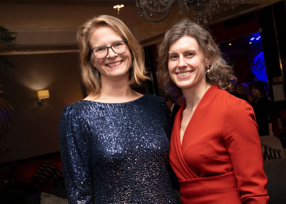 Two people in formal attire, one wearing a sparkly blue dress and one in red, pose together at an indoor event.