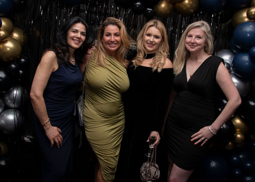 A group of women in evening attire pose together at a festive nighttime event with glowing lights in the background.