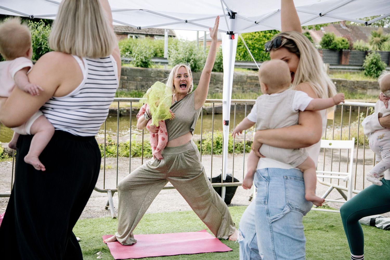 Parent and baby exercise class participants stretching outdoors under white tent.