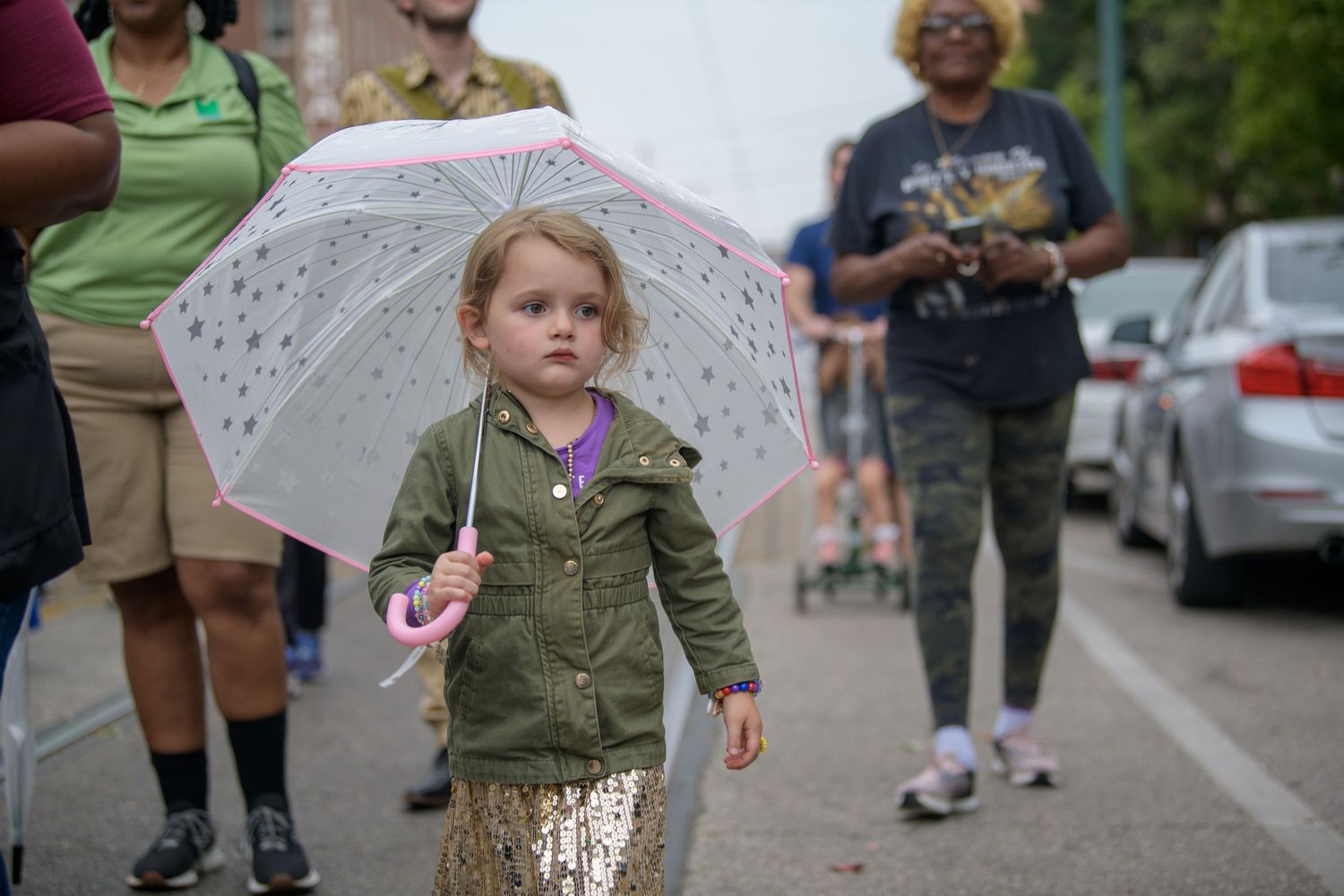 A young person twirls a white lace parasol while walking down a street wearing a green jacket and sparkly pants.