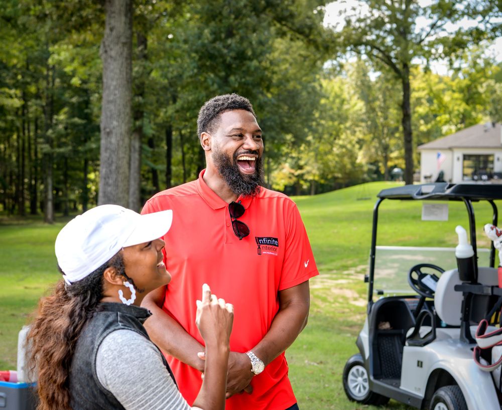 Two people in golf attire stand near a golf cart on a lush green course with trees in the background.