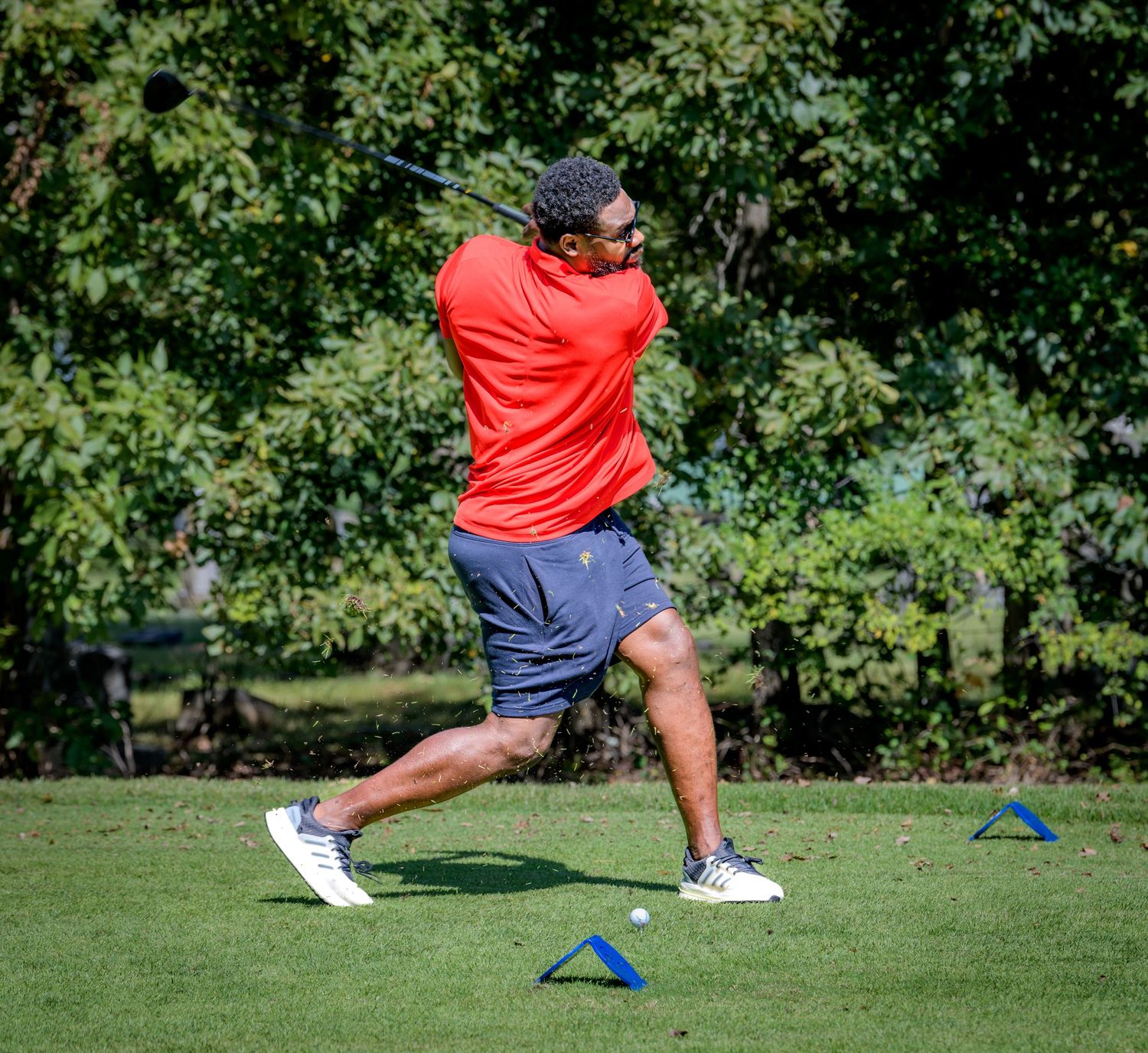 Golfer in red shirt performs swing motion on green grass course with trees in background.