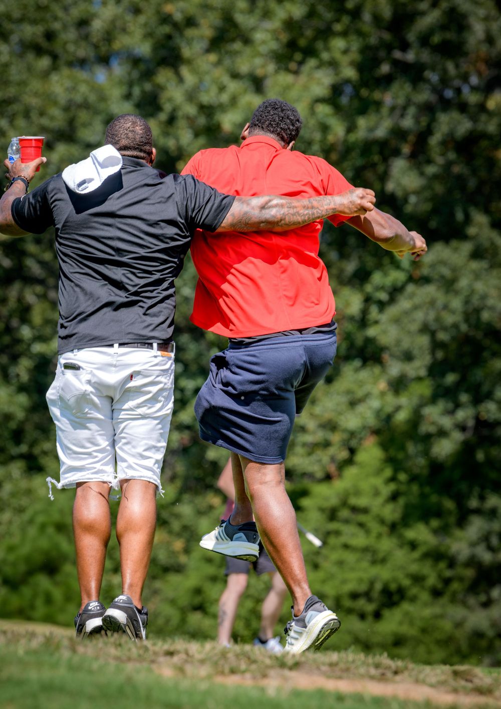Two golfers in casual attire jump and celebrate together on a golf course with green trees in the background.