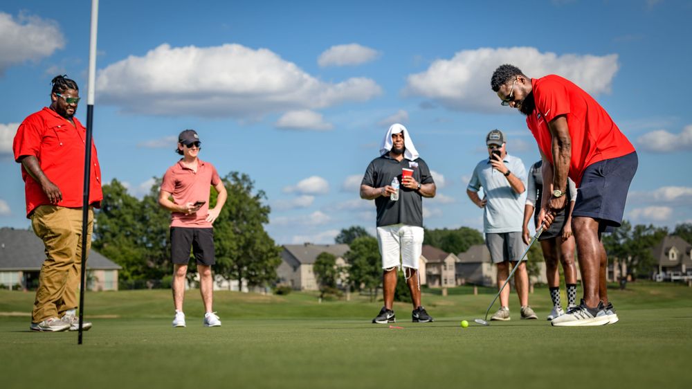 Five people standing on a golf course green, with one person preparing to putt while others observe.