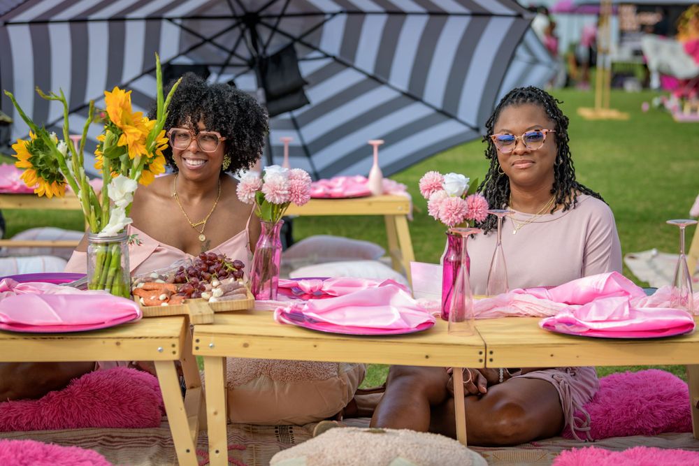 Two guests seated at an outdoor picnic party with pink place settings and striped umbrella at a summer celebration event.