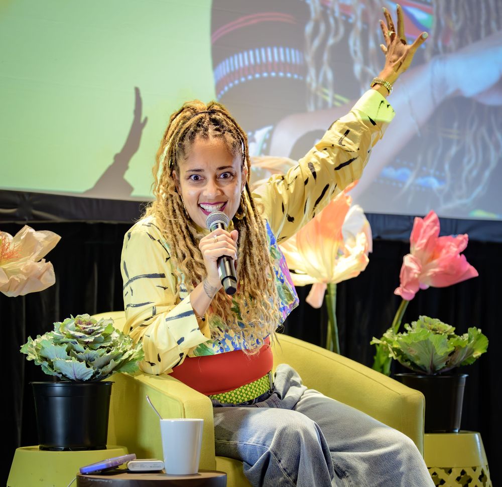 Performer in yellow patterned jacket speaks into microphone on stage with decorative flowers and projected backdrop.