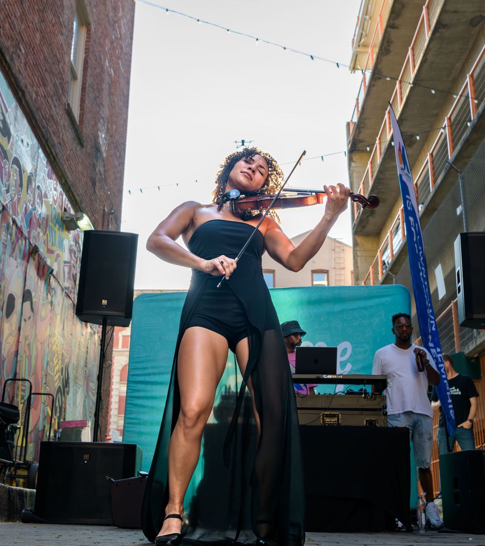 A passionate violinist performs in a black dress with dramatic flair at an outdoor urban venue with speakers and graffiti walls.
