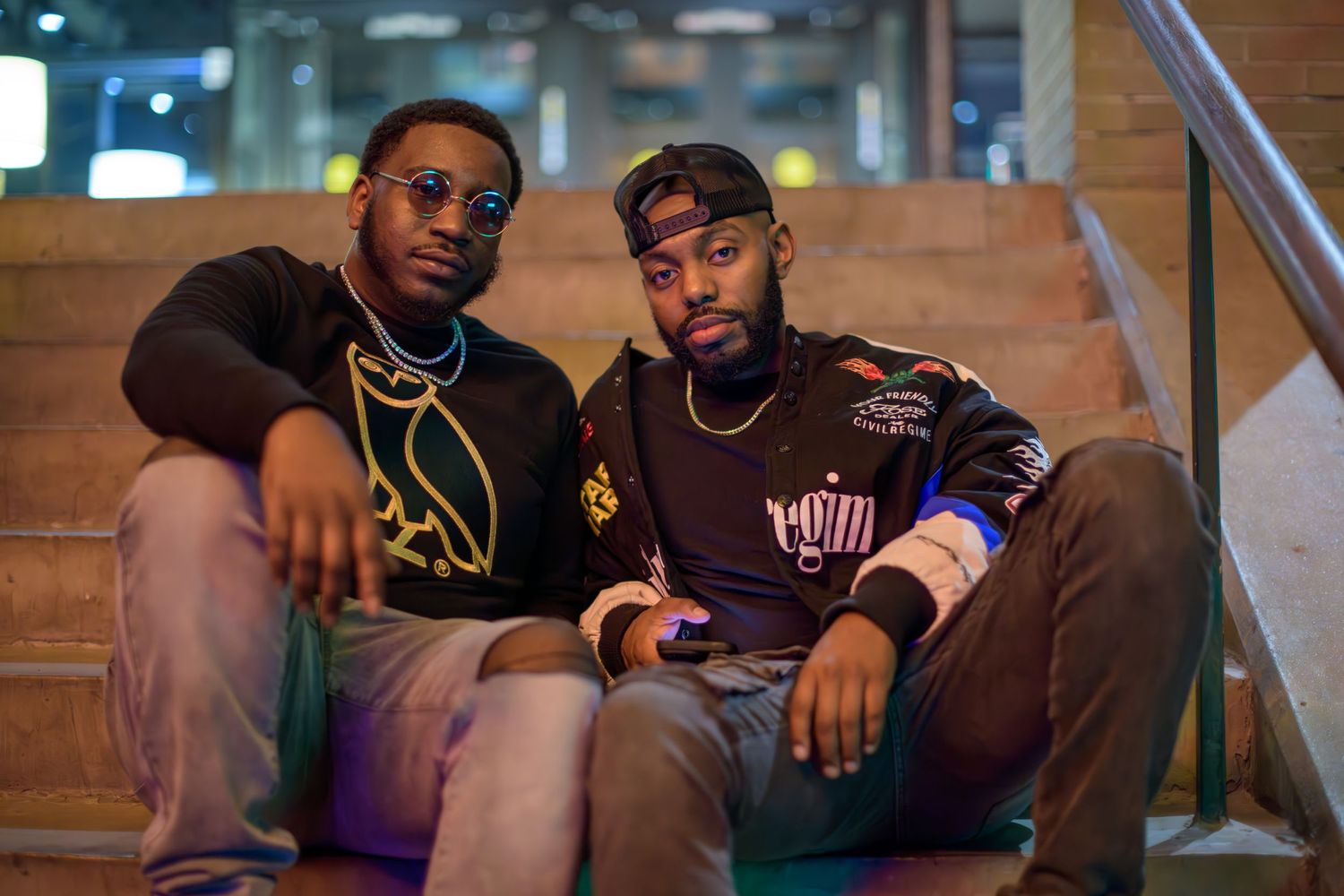 Two young men in casual streetwear sitting on wooden stairs indoors with one wearing sunglasses and gold chain.