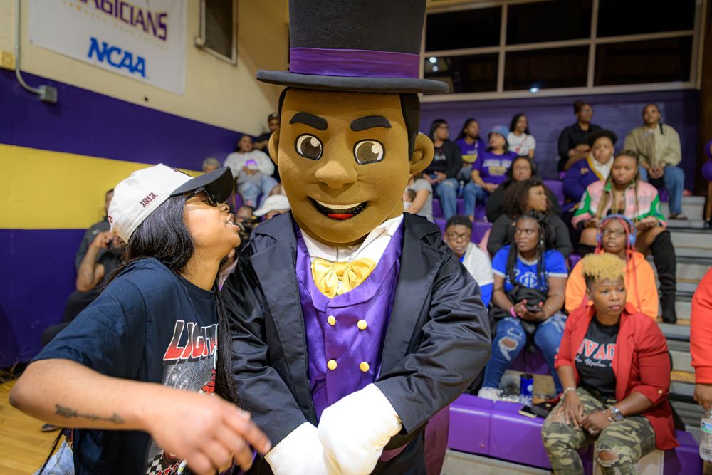Sports mascot in purple outfit and graduation cap interacts with a fan at a basketball arena.