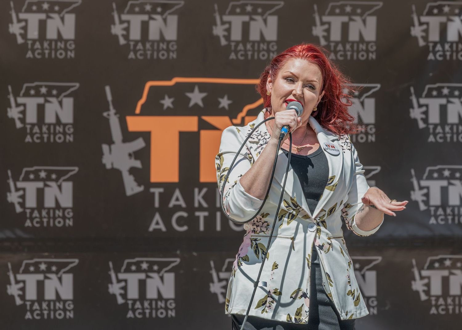 A performer with vibrant red hair sings on stage in a white jacket against a backdrop with Take Action logos.