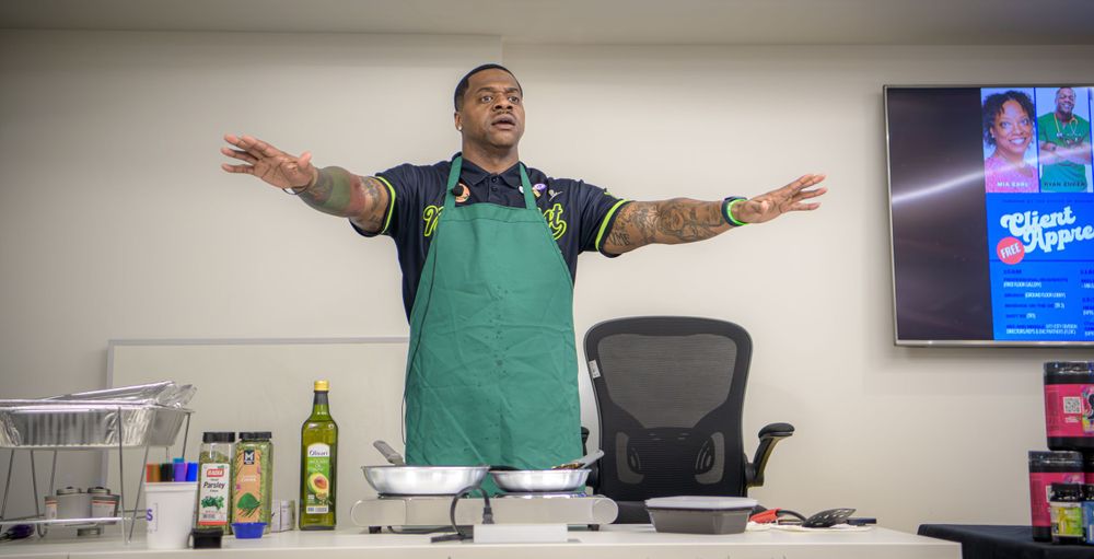 A person wearing a green apron stands with arms outstretched in a kitchen or dining area with bottles on the counter.