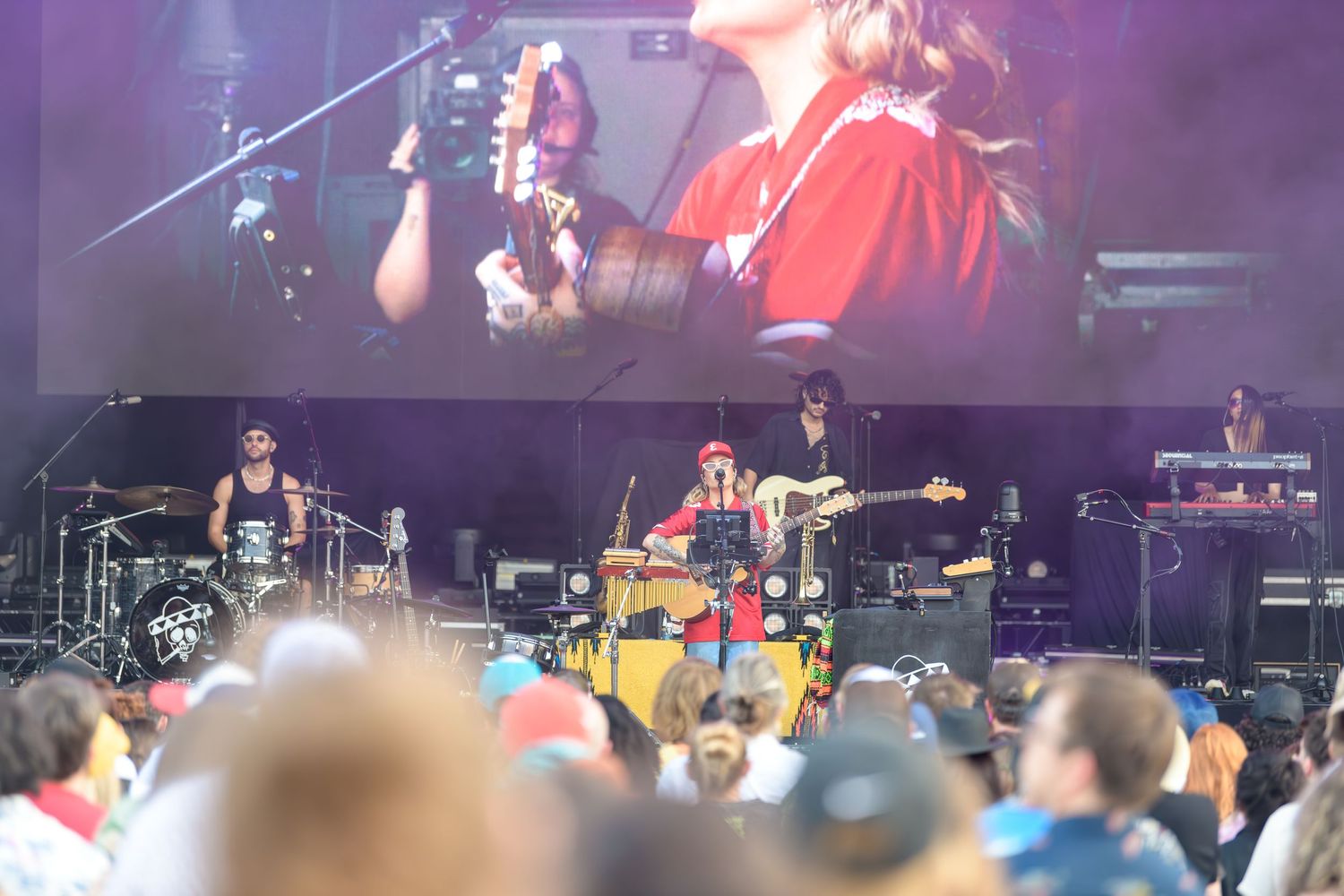 Musicians perform on an outdoor festival stage with a large video screen displaying close-up performance footage above them.