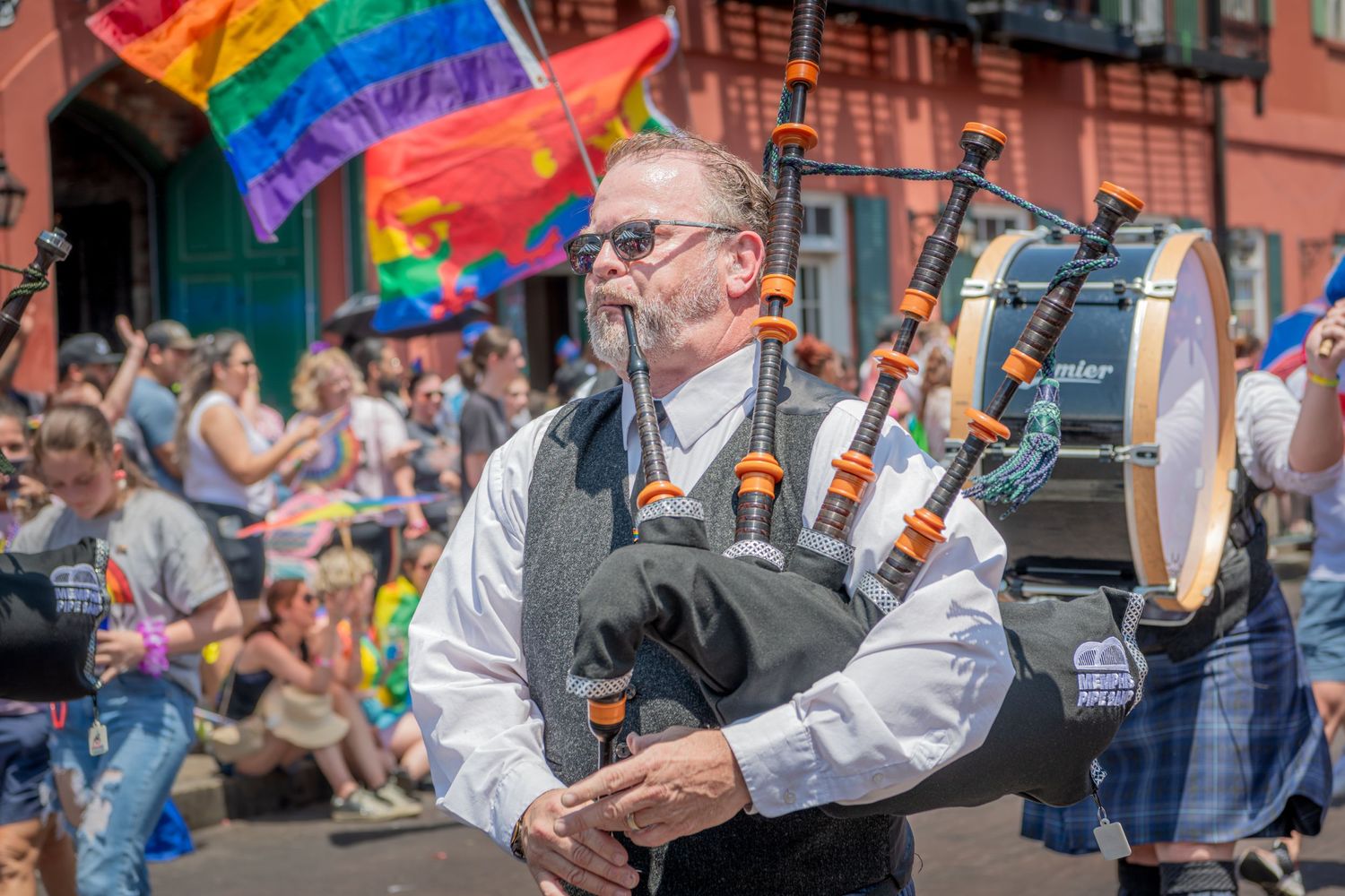 Pride parade participants march with rainbow flag and colorful umbrellas down a city street lined with spectators.
