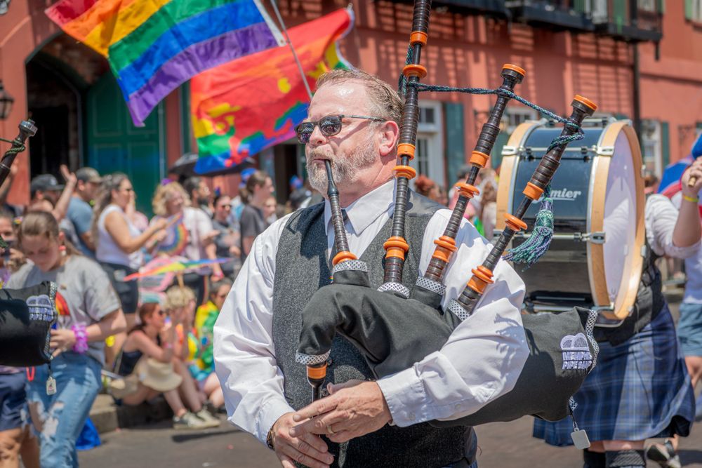 Pride parade participants march with rainbow flag and colorful umbrellas down a city street lined with spectators.