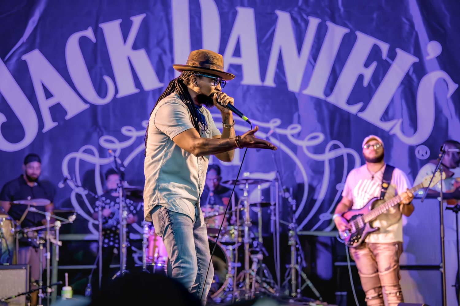 A performer sings on stage with a band in front of a large Jack Daniel's logo during a live concert performance.