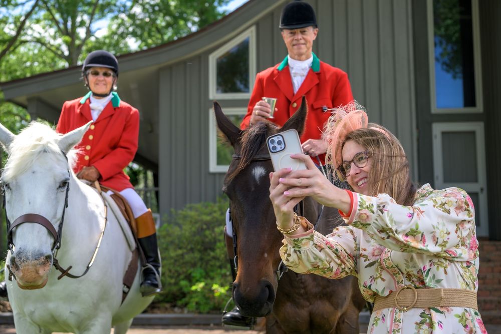 Two people in red riding attire on horses while another takes photos during a fox hunting event.