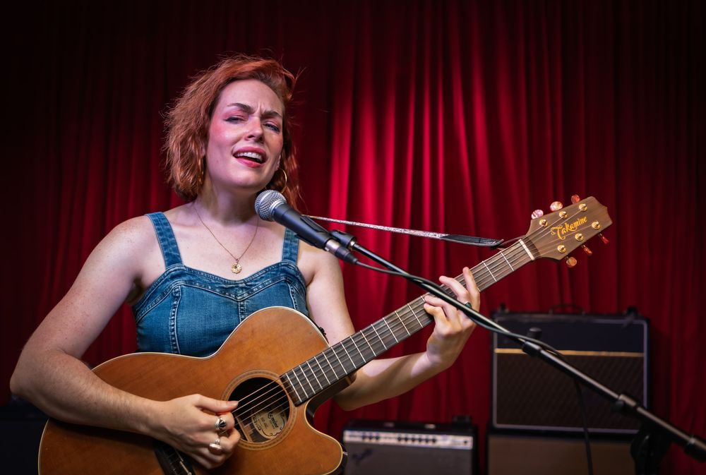 Performer in denim playing acoustic guitar and singing into microphone on stage with red curtain background.
