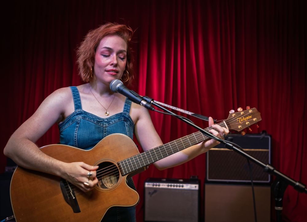 Musician in denim overalls performs with an acoustic guitar against a red curtain backdrop with stage equipment.