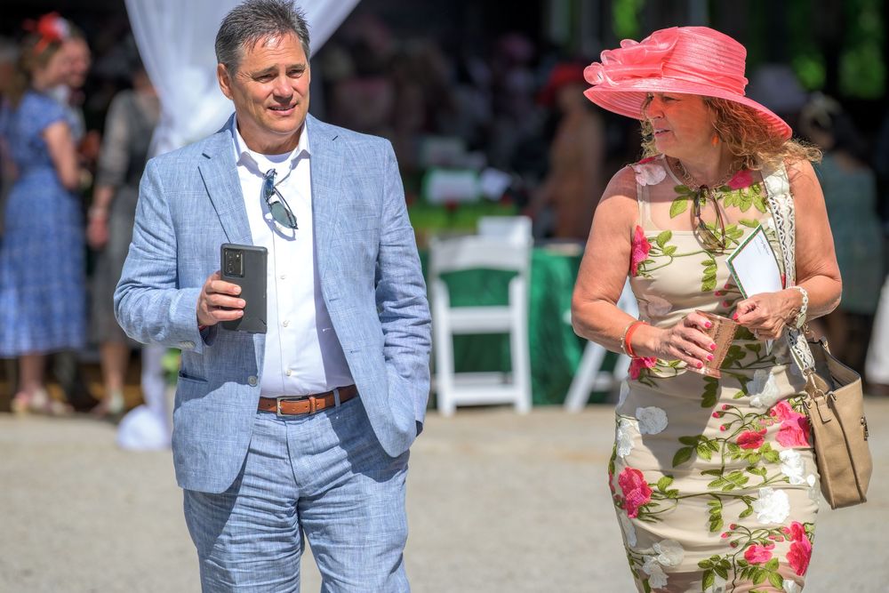 A person in a light blue suit walks alongside someone wearing a floral print dress and wide-brimmed pink hat in a sunny garden.