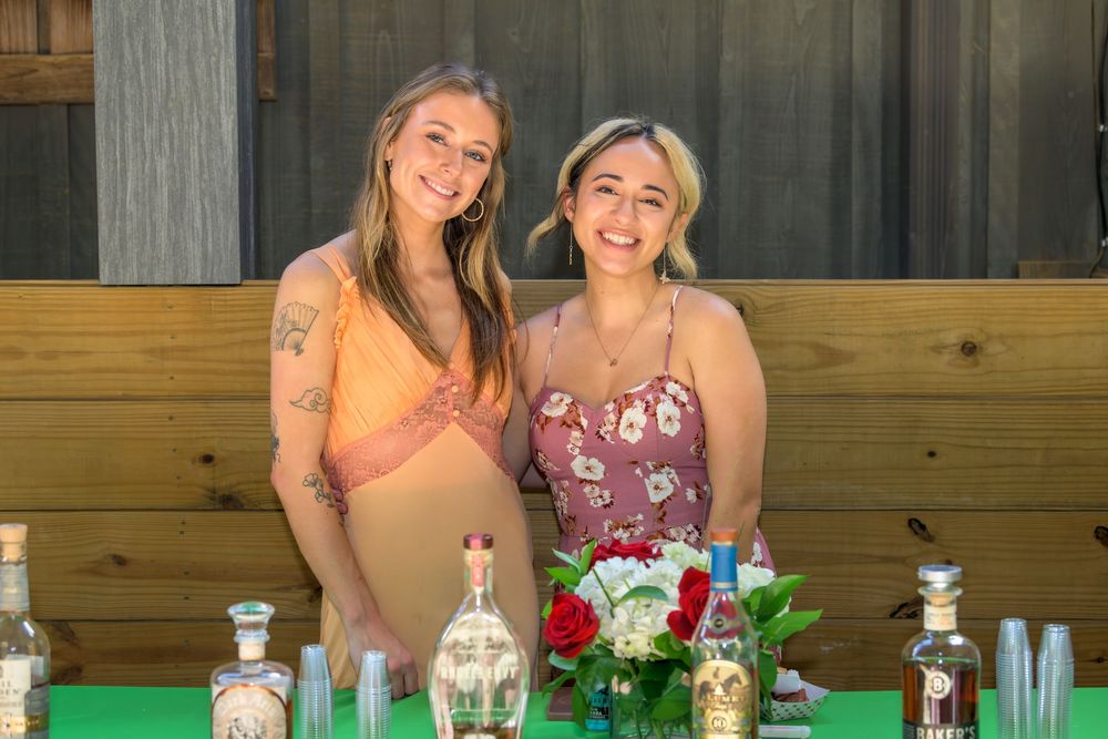 Two friends pose together behind a table with decorative bottles and floral arrangements at an outdoor event.