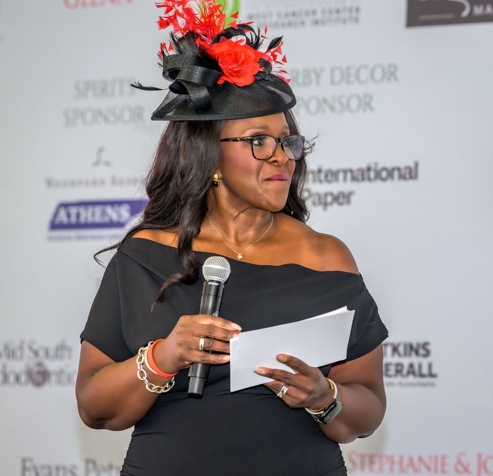 Speaker in black off-shoulder dress and decorative hat with red flowers holds microphone at elegant event backdrop.