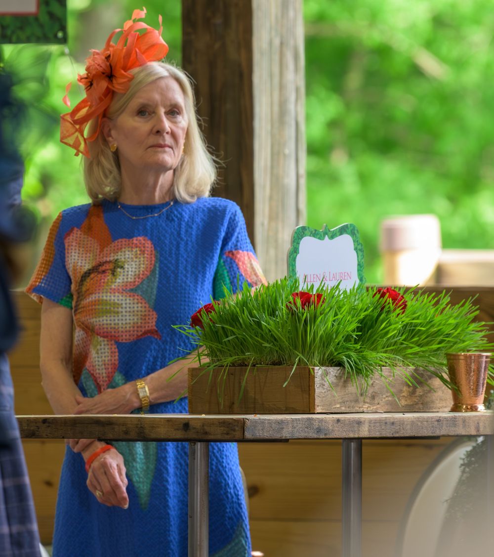 A woman in a blue patterned dress and orange fascinator stands near a decorative grass centerpiece.