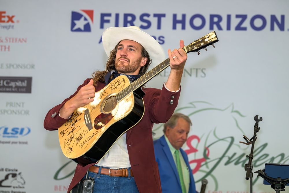 A country music performer in a cowboy hat and burgundy shirt plays an acoustic guitar on stage at a First Horizon sponsored event.