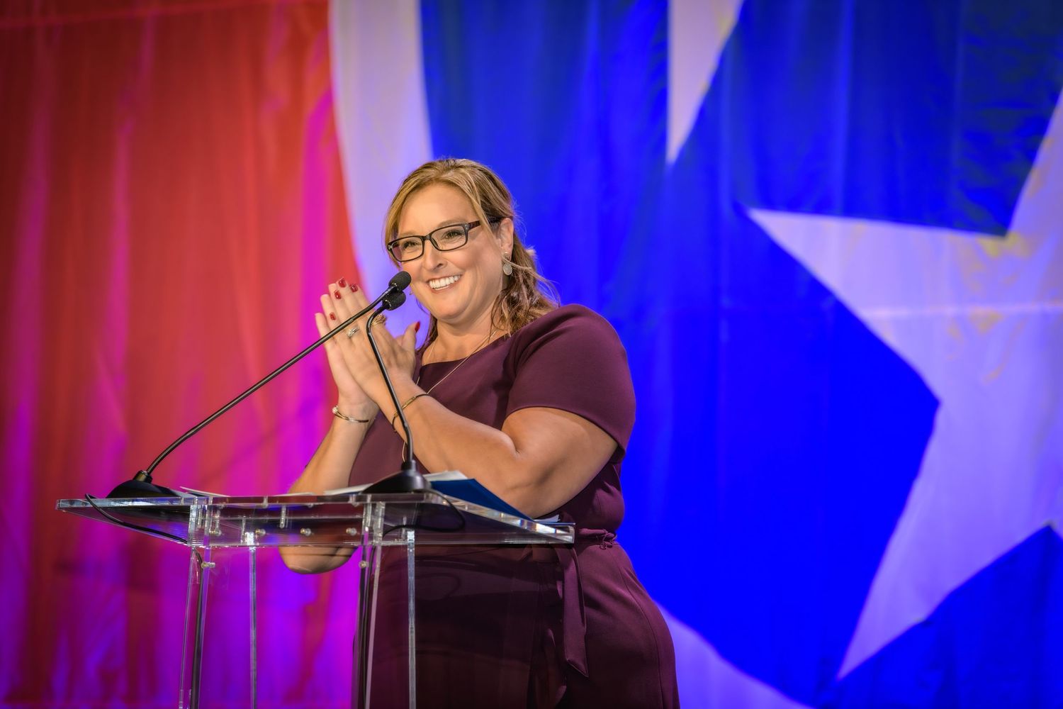 A speaker wearing glasses and dark attire addresses an audience from a podium with red and blue backdrop featuring a star.