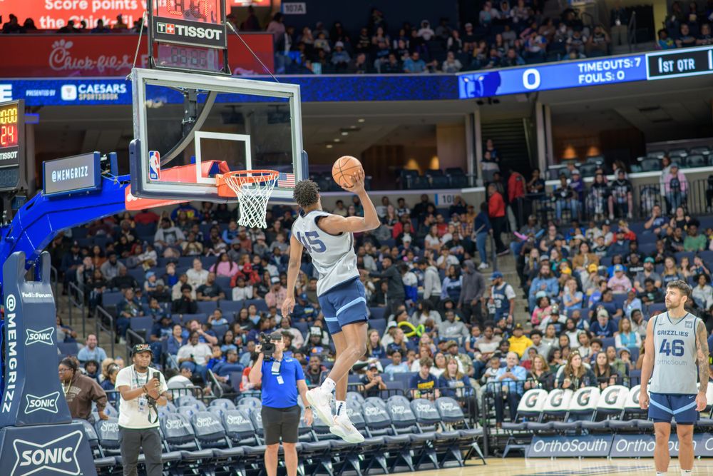 Basketball player soars through the air to complete a dunk during a game at an arena with fans in the background.