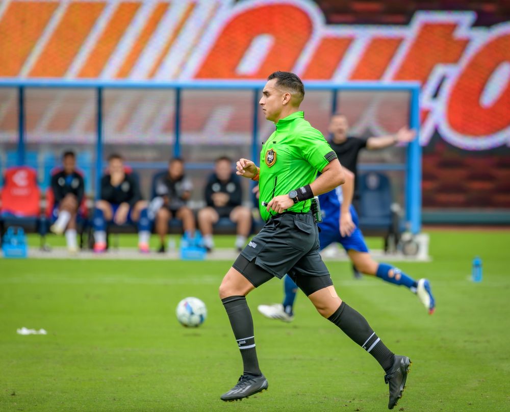 Soccer goalkeeper in neon green jersey makes diving save during match on grass field.