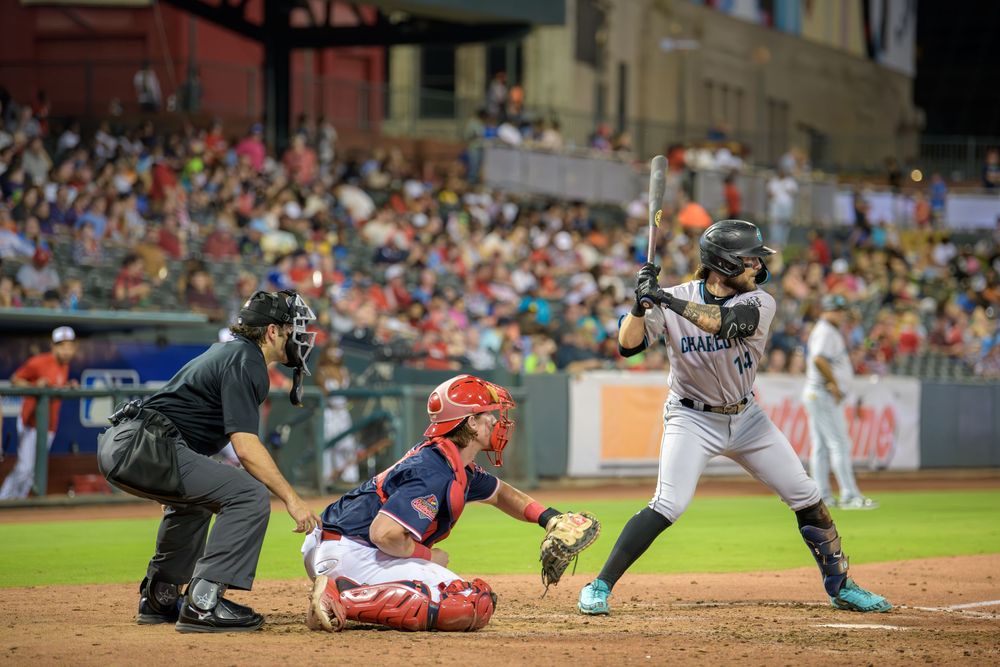 Baseball batter at home plate prepares to swing while catcher and umpire watch during night game at stadium.
