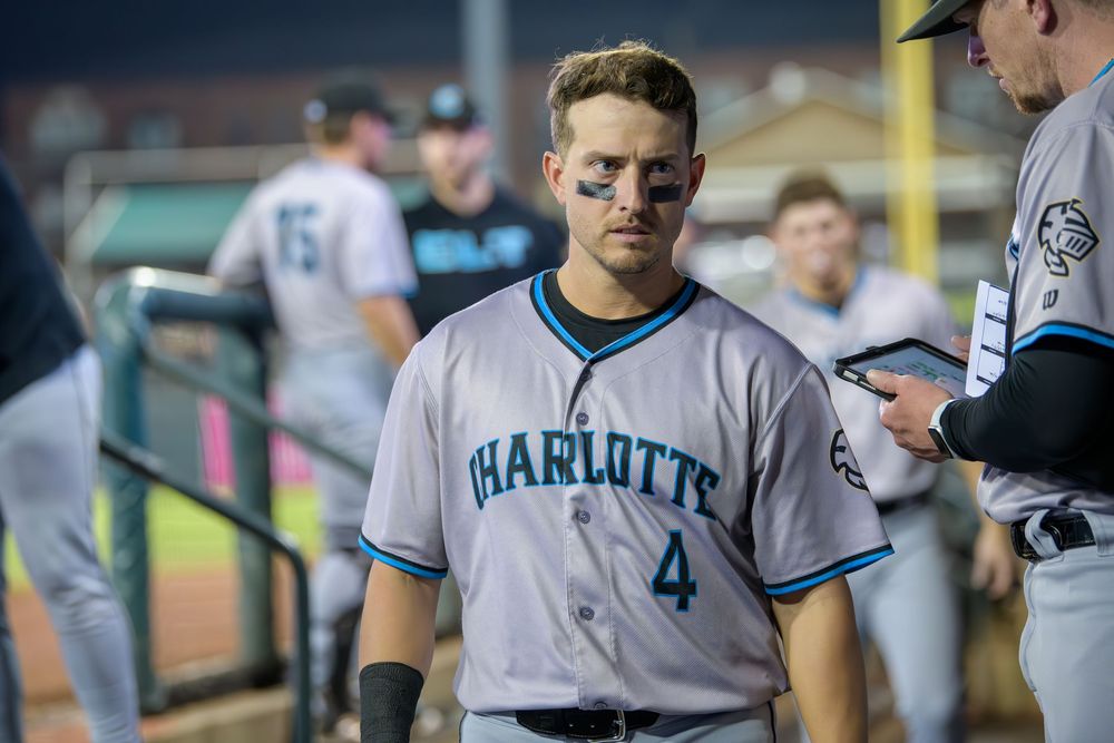 Baseball player in Charlotte uniform stands in dugout during game against Miami Marlins.