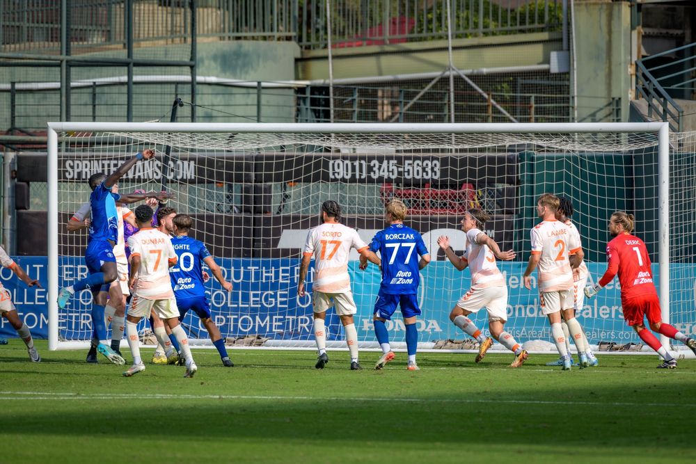 Soccer players compete for the ball during a match on a green field with advertising boards visible in the background.