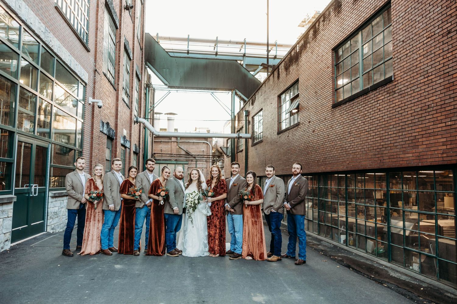 Wedding party in coordinated outfits posed against an industrial brick building backdrop.