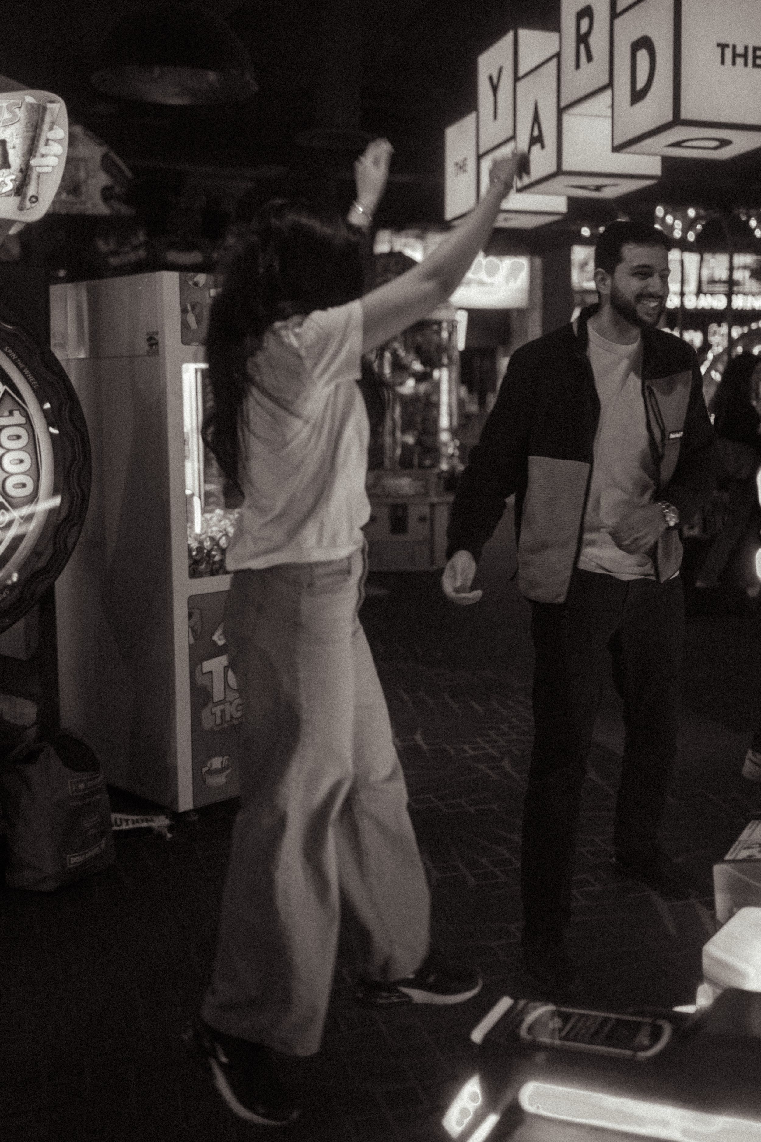 Black and white photo of two people dancing at an arcade bar with YARD sign in background.