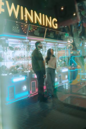 Couple standing in front of neon-lit claw machines under a glowing #WINNING sign in an arcade.