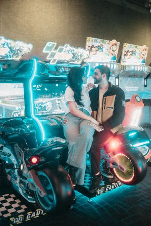Couple sitting together on arcade motorcycle racing game machines with neon lights in a gaming arcade.