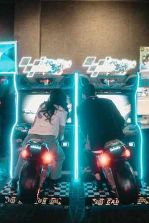 Two players sit on motorcycle arcade game seats with neon blue lighting, competing in a racing game at an arcade.