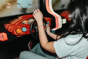 Young girl playing a retro arcade racing game, gripping the steering wheel at an amusement arcade.