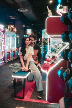 A couple smiles and sits together among glowing neon arcade machines in a vibrant game center.