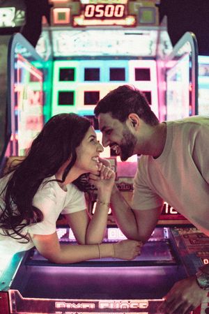 A couple smiles at each other while holding hands at a colorful arcade game machine.