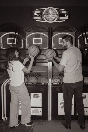 A man and woman hold basketballs at an arcade basketball shooting game in a black and white photo.