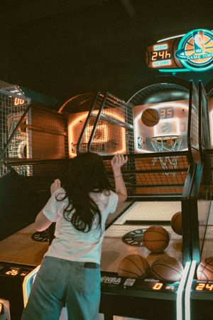 A young girl plays an arcade basketball shooting game, tossing a ball toward the hoop in a dimly lit game room.