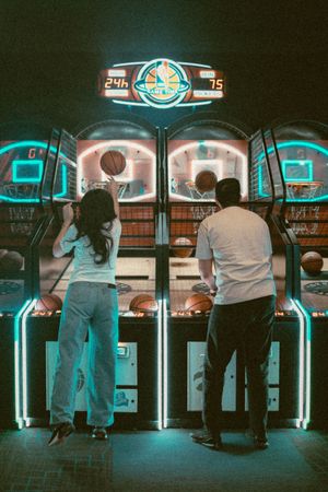 Two people playing basketball arcade game with neon lights in a dark entertainment venue.